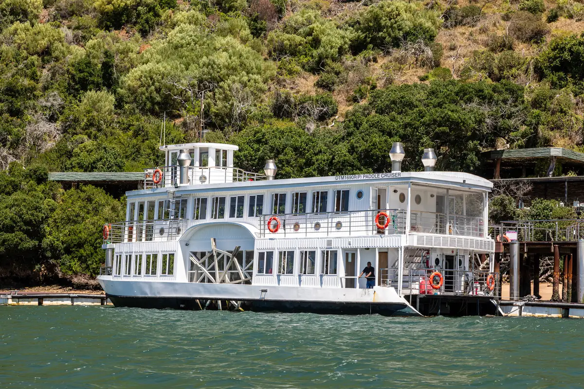 A white double-decker paddle-wheel cruise boat moored at a wooden jetty on green water, with a roofed building on stilts to the right and a steep hillside of coastal bush behind