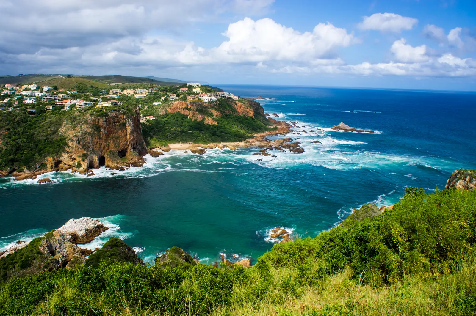 View across a channel of deep blue-green water to a rocky headland with sheer orange-brown cliffs and houses along its top, waves breaking white on rocks at its base, open ocean beyond and green coastal bush in the foreground