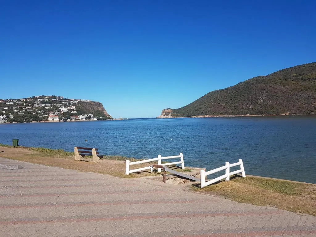 A paved shoreline promenade with a wooden bench and a white-railed slipway running down to calm blue estuary water, with wooded hills and a rocky headland on the far shore under a clear sky