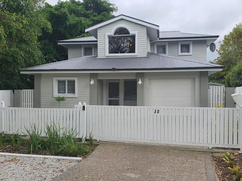 A grey weatherboard double-storey house with a dormer window and a covered entrance, seen from the street behind a white picket fence and a paved brick driveway