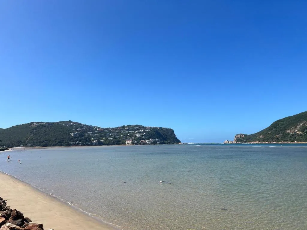 A wide sandy estuary beach with glassy, very shallow water over pale sand in the foreground, houses on a green hillside to the left, and white ocean surf visible breaking through the gap between two headlands in the distance.