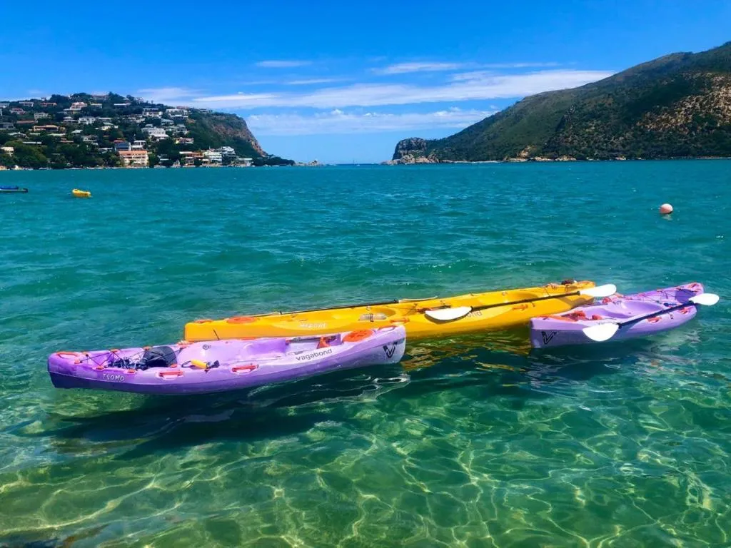 Three kayaks floating on clear turquoise shallow water over a rippled sandy bottom, with wooded hills, houses on a distant hillside and the rocky Knysna Heads in the background