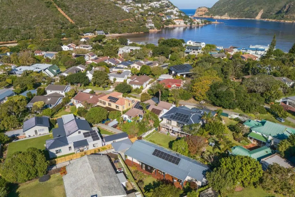 Aerial view over a flat, densely treed residential neighbourhood of detached houses with gardens and solar panels, with calm estuary water and two rocky headlands visible in the distance